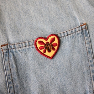 Può includere: Primo piano di una tasca di jeans blu chiaro con una toppa a forma di cuore. La toppa presenta un centro giallo con accenti floreali rossi e un bottone marrone. La tasca è cucita con filo marrone.