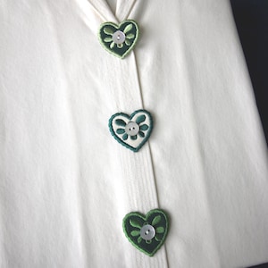 Può includere: Primo piano di un capo color crema con tre applicazioni ricamate verdi a forma di cuore. Ogni applicazione presenta un bottone bianco al centro. Il capo ha una striscia verticale di tessuto al centro.