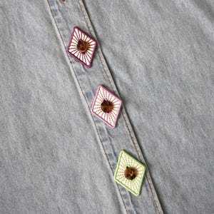 Può includere: Primo piano di una camicia di jeans azzurra con bottoni decorativi a forma di diamante. I bottoni sono marroni con bordi bianchi e colorati ricamati. La camicia ha un classico design con bottoni.