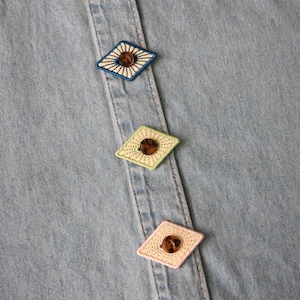 Può includere: Primo piano di una camicia di jeans azzurra con bottoni decorativi a forma di diamante. I bottoni sono ricamati con vari colori, tra cui blu, verde e rosa, e presentano un bottone marrone al centro. La camicia ha un colletto classico e una chiusura con bottoni.