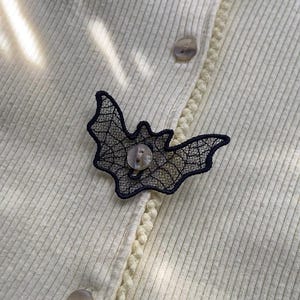 May include: A cream-colored ribbed cardigan featuring a black lace bat-shaped applique attached to the front. The bat applique is centered over one of the translucent buttons, which secures the garment through a vertical placket with a braided trim.