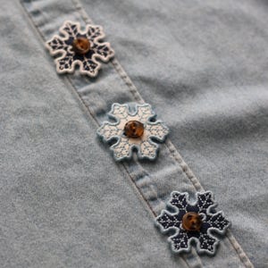 Può includere: Primo piano di una camicia di jeans azzurra con tre bottoni a forma di fiocco di neve. I bottoni hanno un centro marrone e un design ricamato bianco e blu navy. Il tessuto della camicia è leggermente strutturato.