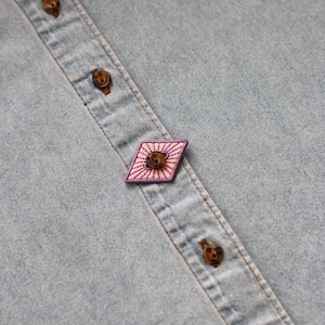Può includere: Primo piano di una camicia di jeans azzurra con bottoni marroni. Una toppa ricamata rosa e viola a forma di diamante è cucita sull'abbottonatura della camicia. La toppa ha un design a raggiera.