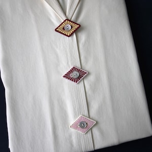 Può includere: Tessuto color crema con tre bottoni decorativi a forma di diamante. Il bottone superiore è bordeaux e giallo, quello centrale è bordeaux e quello inferiore è rosa. Ogni bottone ha un bottone centrale argentato.