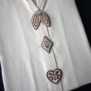 Può includere: Capo color crema con decorativi accenti ricamati marroni. Presenta tre elementi decorativi: un design a forma di foglia, un diamante e un cuore, ciascuno con un bottone centrale. Il tessuto sembra piegato e ha un aspetto strutturato.