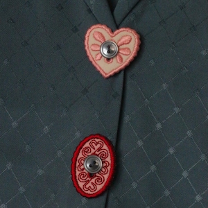 Può includere: Primo piano di un tessuto verde scuro con un motivo a rombi. Sono visibili due bottoni decorativi: un bottone a forma di cuore con ricamo rosa e un bottone ovale rosso con un disegno a spirale. Entrambi hanno bottoni argentati.