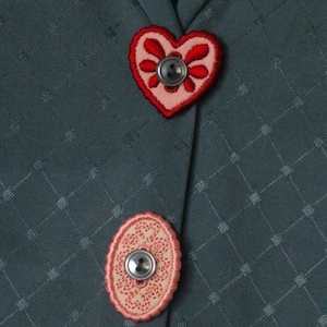 Può includere: Primo piano di un tessuto verde acqua con motivo a rombi. Sono presenti due bottoni decorativi. Il bottone superiore è a forma di cuore con ricami rossi e un bottone grigio. Il bottone inferiore è ovale con ricami rosa e un bottone grigio.