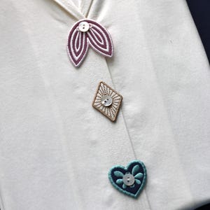 Può includere: Primo piano di un capo bianco con decorazioni ricamate decorative. Presenta un'applicazione a forma di foglia con dettagli viola e bianchi, un'applicazione a forma di diamante e un'applicazione a forma di cuore con un motivo floreale.