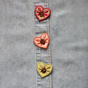 Può includere: Primo piano di una camicia di jeans azzurra con tre toppe ricamate a forma di cuore. Ogni toppa presenta un disegno floreale e un bottone marrone. Le toppe sono nelle tonalità del rosa, arancione e giallo.