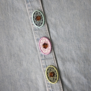 Può includere: Primo piano di una camicia di jeans azzurra con bottoni ovali decorativi. I bottoni sono marroni con bordi ricamati colorati nelle tonalità del verde, rosa e giallo. La camicia ha un classico design con bottoni.