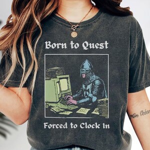 May include: A charcoal gray short-sleeve t-shirt featuring a graphic of a medieval knight in armor sitting at a vintage computer. The text above reads Born to Quest and the text below reads Forced to Clock In, printed in a white serif font.