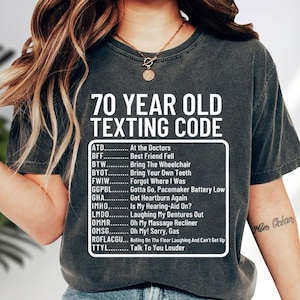 May include: Dark gray t-shirt with the text "70 YEAR OLD TEXTING CODE" and a list of acronyms with their meanings. The shirt has a relaxed fit and a crew neck.