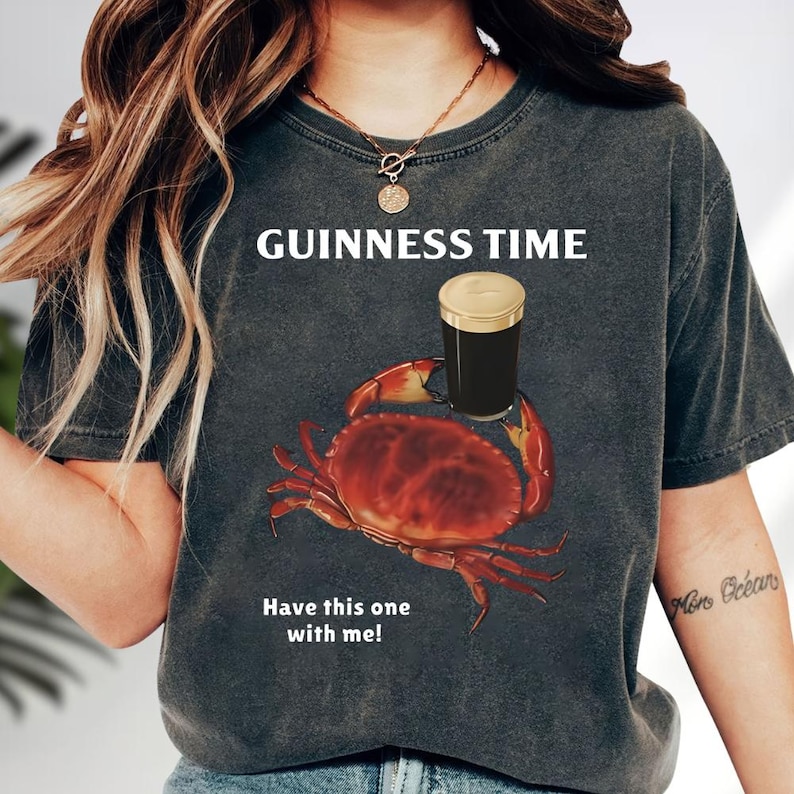 Puede incluir: Camiseta gris oscuro con el texto "GUINNESS TIME" sobre un gr&aacute;fico de un cangrejo rojo sosteniendo una pinta de Guinness. Debajo del cangrejo est&aacute; el texto "Have this one with me!"