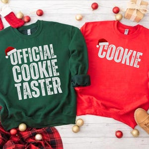 May include: Two crewneck sweatshirts, one green and one red, with holiday-themed text. The green sweatshirt says "OFFICIAL COOKIE TASTER" in white, and the red one says "COOKIE" in white. Both have a Santa hat graphic.