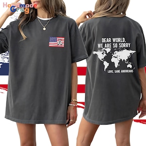 May include: A charcoal grey t-shirt with a small American flag graphic on the front and a larger graphic on the back. The back graphic features a world map with the text "Dear World, We Are So Sorry. Love, Sane Americans".