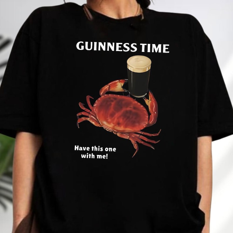 Puede incluir: Camiseta negra con un gr&aacute;fico de un cangrejo sosteniendo una pinta de Guinness. El texto "GUINNESS TIME" est&aacute; encima del cangrejo, y "Have this one with me!" est&aacute; debajo. El cangrejo es rojo y la Guinness es negra y dorada.