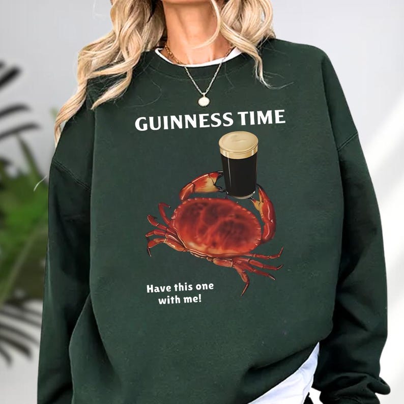 Puede incluir: Sudadera verde oscuro con el texto "GUINNESS TIME" sobre un dibujo de un cangrejo sosteniendo una pinta de Guinness. El cangrejo es rojo y la pinta es negra y marr&oacute;n. El texto "Have this one with me!" est&aacute; debajo.