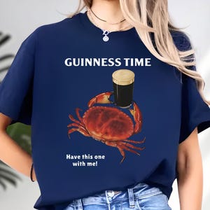 Puede incluir: Camiseta azul marino con el texto "GUINNESS TIME" y un gr&aacute;fico de un cangrejo sosteniendo una pinta de cerveza negra. El texto "Have this one with me!" est&aacute; debajo del cangrejo.