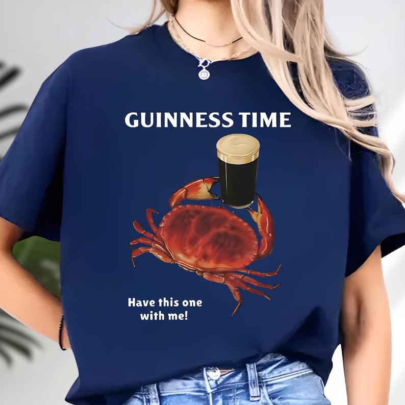 Puede incluir: Camiseta azul marino con el texto "GUINNESS TIME" y un gr&aacute;fico de un cangrejo sosteniendo una pinta de cerveza negra. El texto "Have this one with me!" est&aacute; debajo del cangrejo.