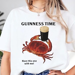 Puede incluir: Camiseta blanca con el texto "GUINNESS TIME" sobre un dibujo de un cangrejo rojo sosteniendo una pinta de Guinness. Debajo est&aacute; el texto "Have this one with me!"