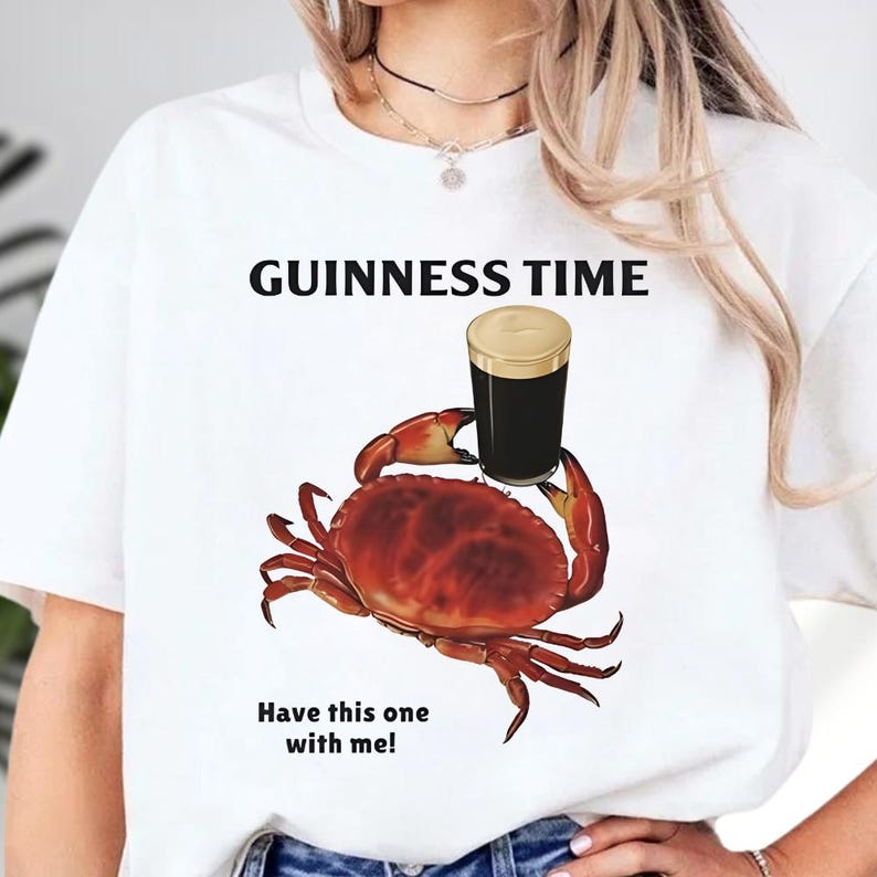Puede incluir: Camiseta blanca con el texto "GUINNESS TIME" sobre un dibujo de un cangrejo rojo sosteniendo una pinta de Guinness. Debajo est&aacute; el texto "Have this one with me!"