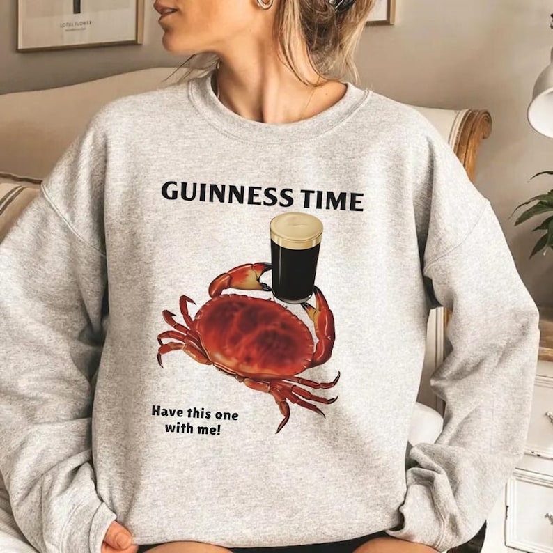 Puede incluir: Una sudadera gris claro con el texto "GUINNESS TIME" sobre un gr&aacute;fico de un cangrejo sosteniendo una pinta de Guinness. Debajo del cangrejo est&aacute; el texto "Have this one with me!"