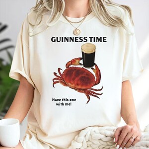 Puede incluir: Camiseta color crema con el texto "GUINNESS TIME" sobre un gr&aacute;fico de un cangrejo sosteniendo una pinta de cerveza negra. El cangrejo es de color rojo-naranja y la cerveza tiene una espuma cremosa. El texto "Have this one with me!" est&aacute; debajo.