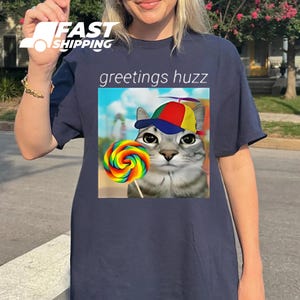 May include: Navy blue t-shirt featuring a graphic of a cat wearing a colorful hat and holding a rainbow lollipop. The text "greetings huzz" is above the graphic. The shirt has a crew neck and short sleeves.