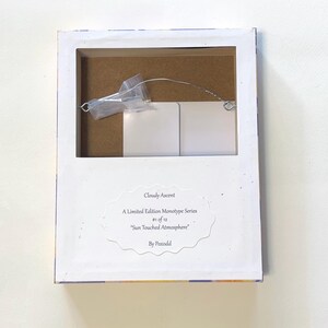May include: The back of a rectangular framed artwork featuring a white paper label with the text: Cloudy Ascent, A Limited Edition Monotype Series, #1 of 12, Sun Touched Atmosphere, By Poxodd. A metal hanging wire and a small plastic bag are attached.