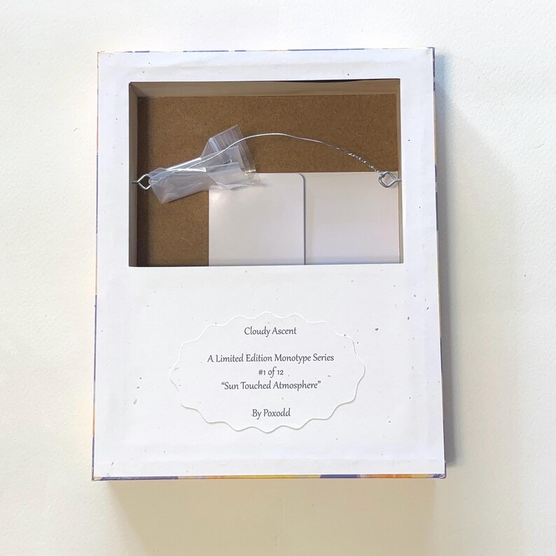 May include: The back of a rectangular framed artwork featuring a white paper label with the text: Cloudy Ascent, A Limited Edition Monotype Series, #1 of 12, Sun Touched Atmosphere, By Poxodd. A metal hanging wire and a small plastic bag are attached.