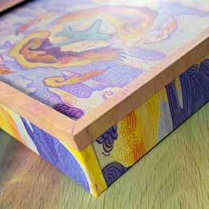 May include: A rectangular box with a light orange frame featuring a colorful illustration on the lid and sides. The artwork depicts a stylized airplane flying through swirling clouds in shades of purple, yellow, and orange with textured line work.
