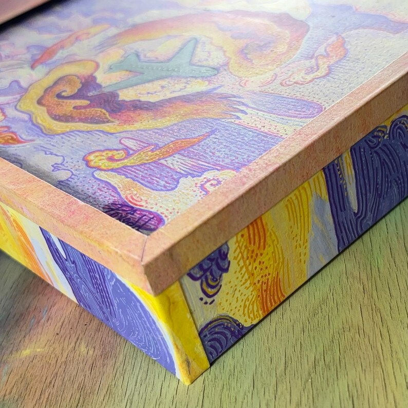 May include: A rectangular box with a light orange frame featuring a colorful illustration on the lid and sides. The artwork depicts a stylized airplane flying through swirling clouds in shades of purple, yellow, and orange with textured line work.
