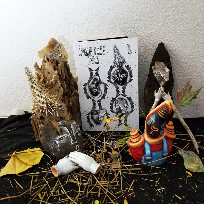 May include: A black and white zine titled "Stone Face Gang" with a number "1" on the cover. The zine is surrounded by various handmade figurines with painted faces, including a figure with a red and blue outfit and a yellow hat.
