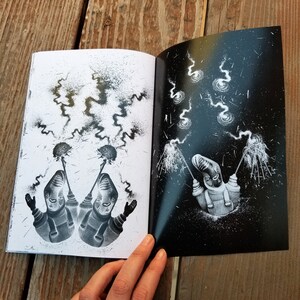 May include: An open book with black and white illustrations. The left page shows two figures with sparklers, while the right page shows a single figure with sparklers against a dark, starry background.