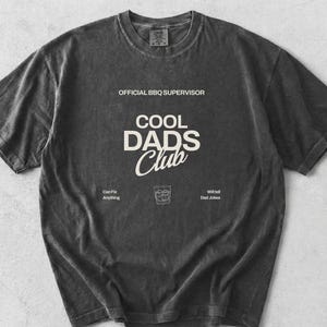 May include: Pepper-colored t-shirt with the text "OFFICIAL BBQ SUPERVISOR" and "COOL DADS Club" in white. Below the text is a graphic of a drink and the phrases "Can Fix Anything" and "Will Tell Dad Jokes".