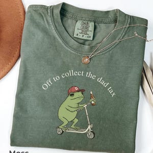 May include: Moss green Comfort Colors t-shirt with a graphic of a frog riding a scooter and holding a beer bottle. The text "Off to collect the dad tax" is arched above the frog. A gold necklace is draped on the shirt.