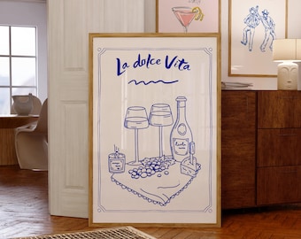 May include: A framed art print with a blue and white illustration of a wine scene, including wine glasses, a bottle, grapes, and cheese. The words "La dolce Vita" are at the top. Two other framed prints are visible in the background.