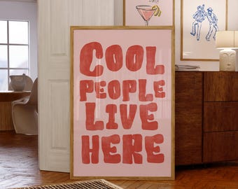 May include: A rectangular wall art print in a light wooden frame featuring the red text COOL PEOPLE LIVE HERE in a bold, wavy font against a light pink background. The print is displayed upright on a wooden floor.