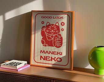 May include: Framed print featuring a red Maneki Neko cat illustration with the words "GOOD LUCK" and "MANEKI NEKO" in red. The print is in a wooden frame and is displayed on a wooden cabinet. A green vase is on the right.
