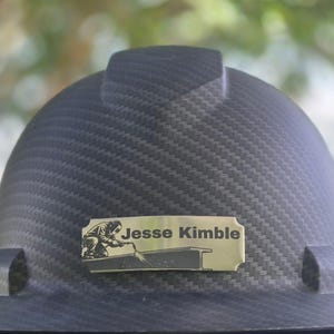 May include: A black carbon fiber pattern hard hat featuring a rectangular gold-toned metal nameplate centered on the front. The nameplate displays the text Jesse Kimble alongside a black graphic illustration of a person welding a steel beam.