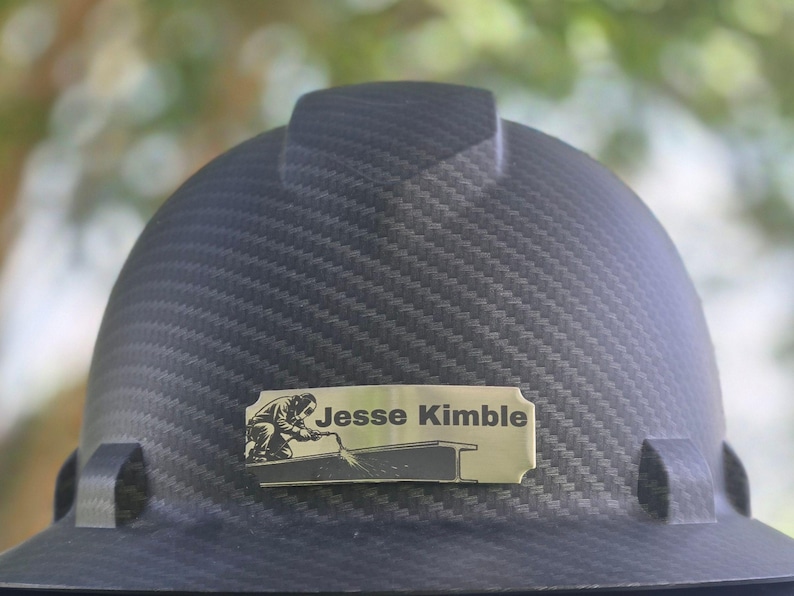 May include: A black carbon fiber pattern hard hat featuring a rectangular gold-toned metal nameplate centered on the front. The nameplate displays the text Jesse Kimble alongside a black graphic illustration of a person welding a steel beam.