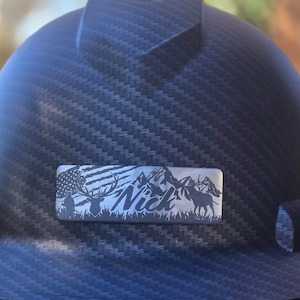 May include: A black hard hat with a textured surface. A rectangular metal emblem features an American flag, mountains, deer, and the name "Nick". The hat is designed for construction or industrial use.