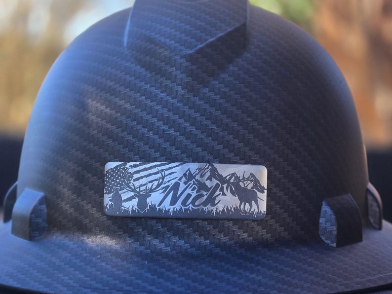 May include: A black hard hat with a textured surface. A rectangular metal emblem features an American flag, mountains, deer, and the name "Nick". The hat is designed for construction or industrial use.