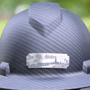 May include: A dark grey hard hat with a carbon fibre texture pattern. A rectangular silver-toned metal plate is attached to the front, featuring the text "Lisa McAllister" above an illustration of a semi-truck and an oil derrick pump jack.