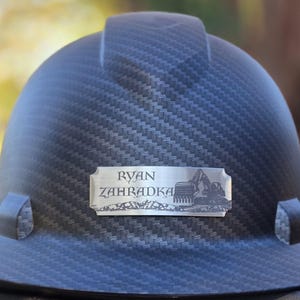 May include: A dark gray hard hat with a textured pattern. A silver nameplate displays the name "RYAN ZAHADKA" and an excavator graphic. The hat is designed for construction or industrial use.