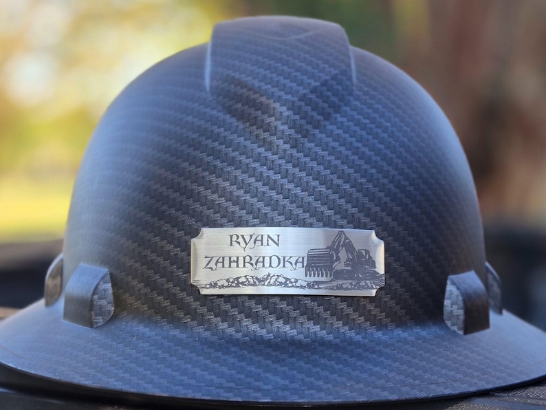 May include: A dark gray hard hat with a textured pattern. A silver nameplate displays the name "RYAN ZAHADKA" and an excavator graphic. The hat is designed for construction or industrial use.