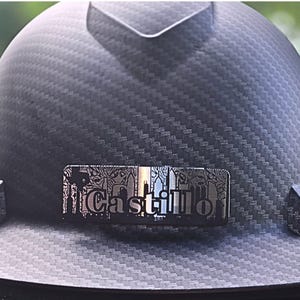 May include: A black hard hat with a textured carbon fiber pattern. A silver nameplate with the name "Castillo" is affixed to the front. The nameplate has a decorative border and a cityscape design.