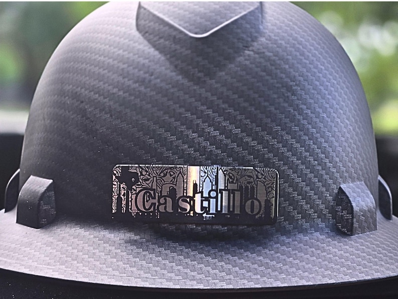 May include: A black hard hat with a textured carbon fiber pattern. A silver nameplate with the name "Castillo" is affixed to the front. The nameplate has a decorative border and a cityscape design.