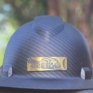 May include: A black hard hat with a textured carbon fiber pattern. A gold-colored rectangular emblem features a train illustration and the text "CARNEBECK". The hat has a brim and a top ridge.