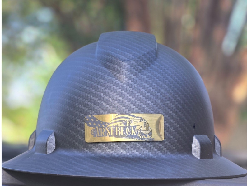 May include: A black hard hat with a textured carbon fiber pattern. A gold-colored rectangular emblem features a train illustration and the text "CARNEBECK". The hat has a brim and a top ridge.